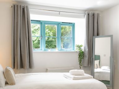 A bedroom with a bed, folded towels, a window with blue frames and grey curtains, and a standing mirror with plants near Newlyn in Mawnan Smith Near Falmouth