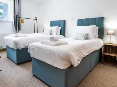 A bedroom with two single beds white bedding and blue headboards near a window and a wooden bedside table with a lamp at Newlyn in Mawnan Smith Near Falmouth