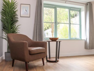 A brown armchair next to a small round wooden table with a bowl and a large window with grey curtains and a tall green plant at Penpoll in Falmouth