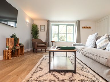 A living room with a grey sofa and cushions a brown armchair wooden crates with plants a TV on the wall and a coffee table on a rug at Penpoll in Falmouth
