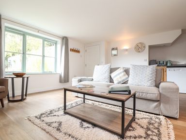 A living room with a sofa, coffee table, side table, large window, wall clock, and kitchen appliances at Penpoll in Falmouth