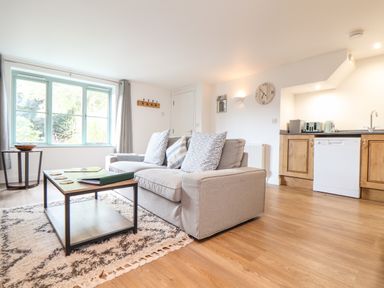 A living room with a grey sofa and patterned cushions next to a wooden coffee table and a kitchen area at Penpoll in Falmouth
