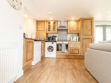 A kitchen with wooden cabinets, a washing machine, dishwasher, oven, microwave, and a gray sofa nearby at Penpoll in Falmouth