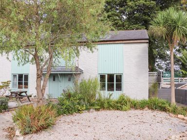 Exterior view of a white stone house with light green doors and windows surrounded by trees bushes and gravel at Penpoll in Falmouth