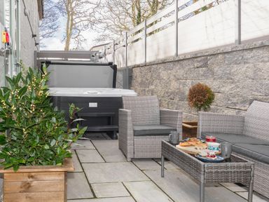 An outdoor patio area with a hot tub a wicker sofa and armchair a glass coffee table with food and drinks and plants in wooden pots at Penpoll in Falmouth