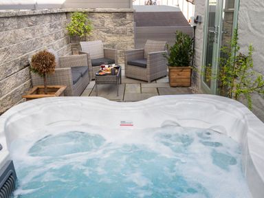 An outdoor patio area with a hot tub in the foreground wicker chairs and a table with food and drinks against a stone wall at Penpoll in Falmouth