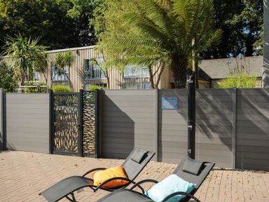 An outdoor patio with two black lounge chairs with orange and blue pillows and a gray privacy fence with a gate and plants behind it at Porthallow in Mawnan Smith Near Falmouth