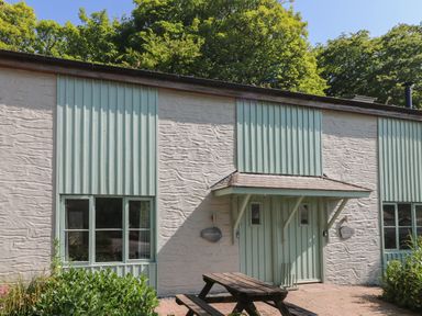 The exterior of a building with textured white walls and pale green vertical panels with a wooden picnic table in front surrounded by plants at Porthallow in Mawnan Smith Near Falmouth