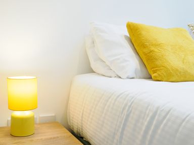 A bed with white bedding and pillows and a yellow cushion next to a wooden bedside table with a yellow lamp