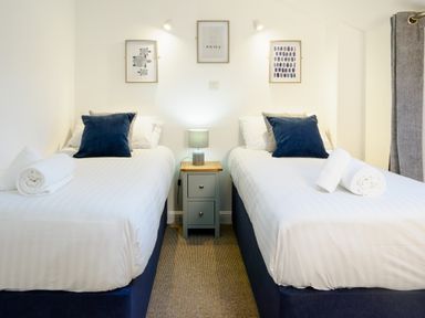A bedroom with two single beds with white bedding and blue pillows a nightstand with a lamp between the beds three framed pictures on the wall and towels on the bed at Porthallow in Mawnan Smith Near Falmouth