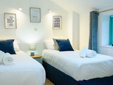 A bedroom with two single beds with blue pillows and rolled white towels on them a nightstand with a lamp and framed pictures on the wall at Porthallow in Mawnan Smith Near Falmouth