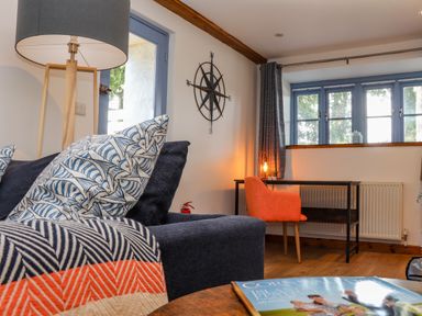 A living room with a blue sofa with patterned pillows a wooden floor lamp a desk with an orange chair and blue framed windows at St Keverne in Mawnan Smith Near Falmouth