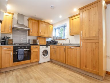 A kitchen with wooden cabinets, built-in oven, washing machine, microwave, toaster, kettle, sink, and window at St Keverne in Mawnan Smith Near Falmouth