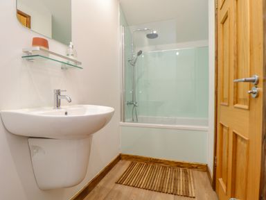 A bathroom with a wall-mounted sink a glass shelf a bathtub with a glass shower screen and wooden door at St Keverne in Mawnan Smith Near Falmouth