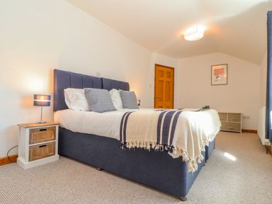 A bedroom with a blue bed, bedside table with lamp, a wooden door, drawer unit, and framed picture on the wall at St Keverne in Mawnan Smith Near Falmouth