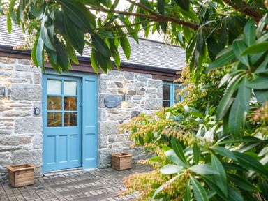 A stone house with a blue door and window framed by green leafy plants at Manacles in Mawnan Smith Near Falmouth