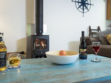A living room with a wood burning stove a blue table with drinks a fruit bowl a brown sofa and a wall clock at Manacles in Mawnan Smith Near Falmouth