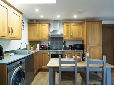 A kitchen with wooden cabinets, a dining table with chairs, washing machine, dishwasher, oven, and kitchen appliances at Manacles in Mawnan Smith near Falmouth