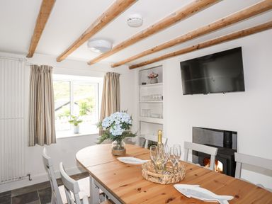 A dining room with a wooden table and white chairs a TV mounted on the wall and a fireplace with fire burning at Isfryn in Llanengan near Abersoch