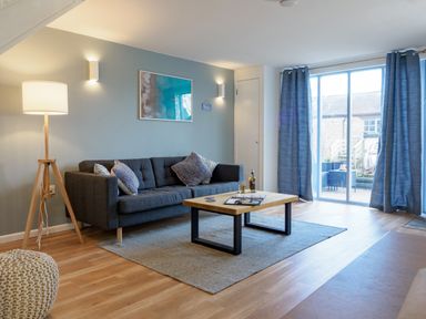 A living room with a dark sofa, coffee table, floor lamp, and sliding glass door with curtains at Coverack in Mawnan Smith Near Falmouth