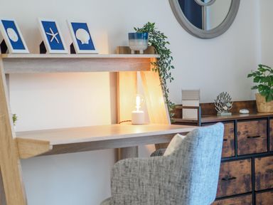 A wooden desk with framed sea-themed decorations a small lamp and a plant next to a gray chair and a wooden cabinet with plants at Coverack in Mawnan Smith Near Falmouth