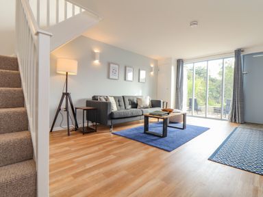 A living room with a gray sofa a wood and metal coffee table a blue rug a floor lamp stairs and sliding glass doors at Helford in Mawnan Smith Near Falmouth