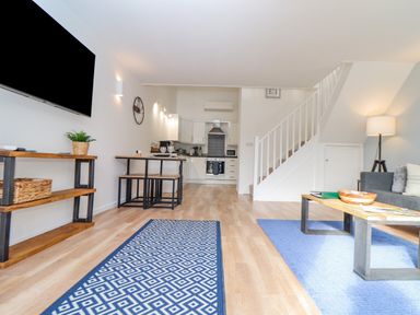 A living room with a wall-mounted television and a wooden shelf a dining table with benches a staircase and a kitchen in the background at Helford in Mawnan Smith Near Falmouth
