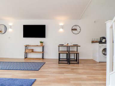 A living room with a wall mounted tv a wooden table a clock two stools a washing machine and coffee maker at Helford in Mawnan Smith Near Falmouth