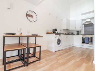 A kitchen with white cabinets a washing machine a dining table with stools and a wall clock at Helford in Mawnan Smith Near Falmouth