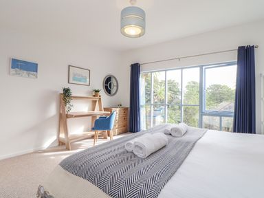 A bedroom with a bed and rolled towels a wooden desk with a blue chair a chest of drawers and large windows with blue curtains at Helford in Mawnan Smith Near Falmouth
