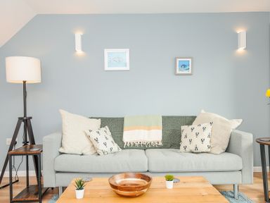 A living room with a grey sofa with cushions and a throw blanket a wooden coffee table with plants a floor lamp and framed pictures on a blue wall at Mousehole in Mawnan Smith Near Falmouth