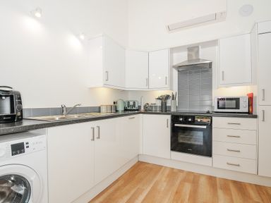 A kitchen with white cabinets a black countertop a washing machine a coffee maker a toaster a kettle an oven a microwave and wooden floor at Mousehole in Mawnan Smith Near Falmouth