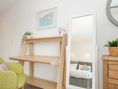 A wooden desk next to a green chair with a striped cushion and a full-length white framed mirror reflecting a bed in a bedroom