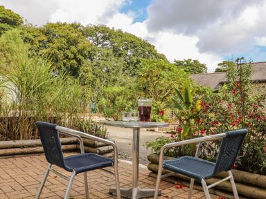 An outdoor patio with a round metal table and two blue woven chairs surrounded by plants and flowers at Sennen in Mawnan Smith Near Falmouth