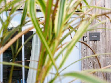 A wooden exterior wall with a glass door a curtain a doorbell and a sign that says Sennen partially obscured by green plants at Sennen in Mawnan Smith Near Falmouth