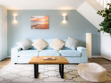 A living room with a light blue sofa cushions a wooden coffee table a beige textured pouf a plant and a sunset beach wall art at Sennen in Mawnan Smith Near Falmouth