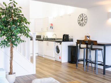 A kitchen area with white cabinets a washing machine a microwave a coffee machine and a table with two stools at Sennen Mawnan Smith Near Falmouth