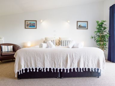 A bedroom with a bed with white bedding and rolled towels two framed pictures on the wall a brown armchair and a tall green plant at Sennen in Mawnan Smith Near Falmouth