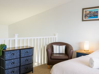 A bedroom corner with a brown armchair a chest of drawers a bed with a rolled towel and a bedside table with a lamp at Sennen in Mawnan Smith Near Falmouth