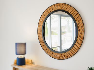 A round wall mirror with a woven frame above a wooden shelf with a blue table lamp