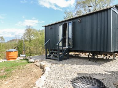 A small black corrugated metal cabin on wheels in a gravel yard with steps and an outdoor wooden hot tub at Coraldie Laggan near Newtonmore