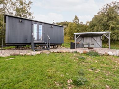 A small grey cabin with stairs and wheels next to a covered seating area with two chairs on grass at Coraldie in Laggan near Newtonmore