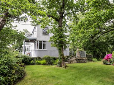 A house with a balcony surrounded by green trees and a grassy lawn at Ynys Castell in Menai Bridge