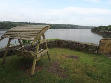 A wooden bench swing with a slatted roof on a grassy area overlooking a body of water with boats and trees at Ynys Castell in Menai Bridge