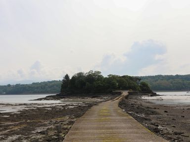 A concrete causeway leading to a small island with trees surrounded by water and rocky shore at Ynys Castell in Menai Bridge