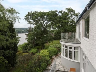 A curved house with a balcony and large windows overlooking a garden with trees and a river in the background at Ynys Castell in Menai Bridge