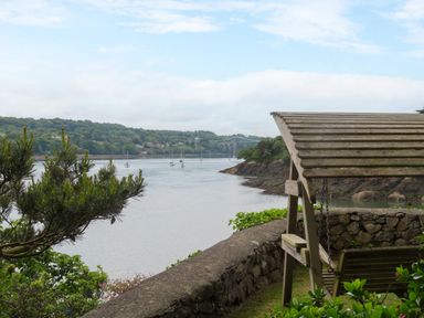 A riverside view with a wooden swing seat under a pergola near a stone wall and boats on the water at Ynys Castell in Menai Bridge