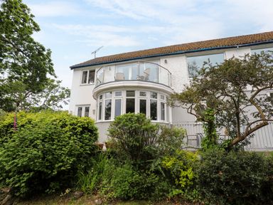 A two-story white house with a balcony and large windows surrounded by green bushes and trees at Ynys Castell in Menai Bridge