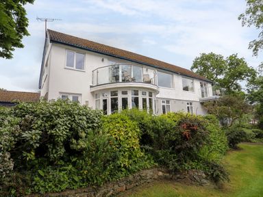 A two-story white house with balconies and windows surrounded by green bushes and trees at Ynys Castell in Menai Bridge