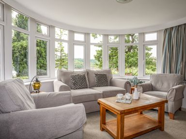 A living room with a sofa, two armchairs, a wooden coffee table, and large windows showing greenery at Ynys Castell in Menai Bridge